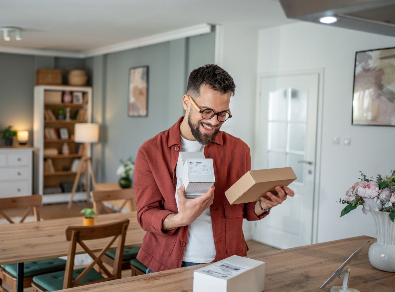 A man with glasses smiles while holding a small package in a cozy, well-lit living room. The room features wooden furniture and decorative plants, creating a warm atmosphere.