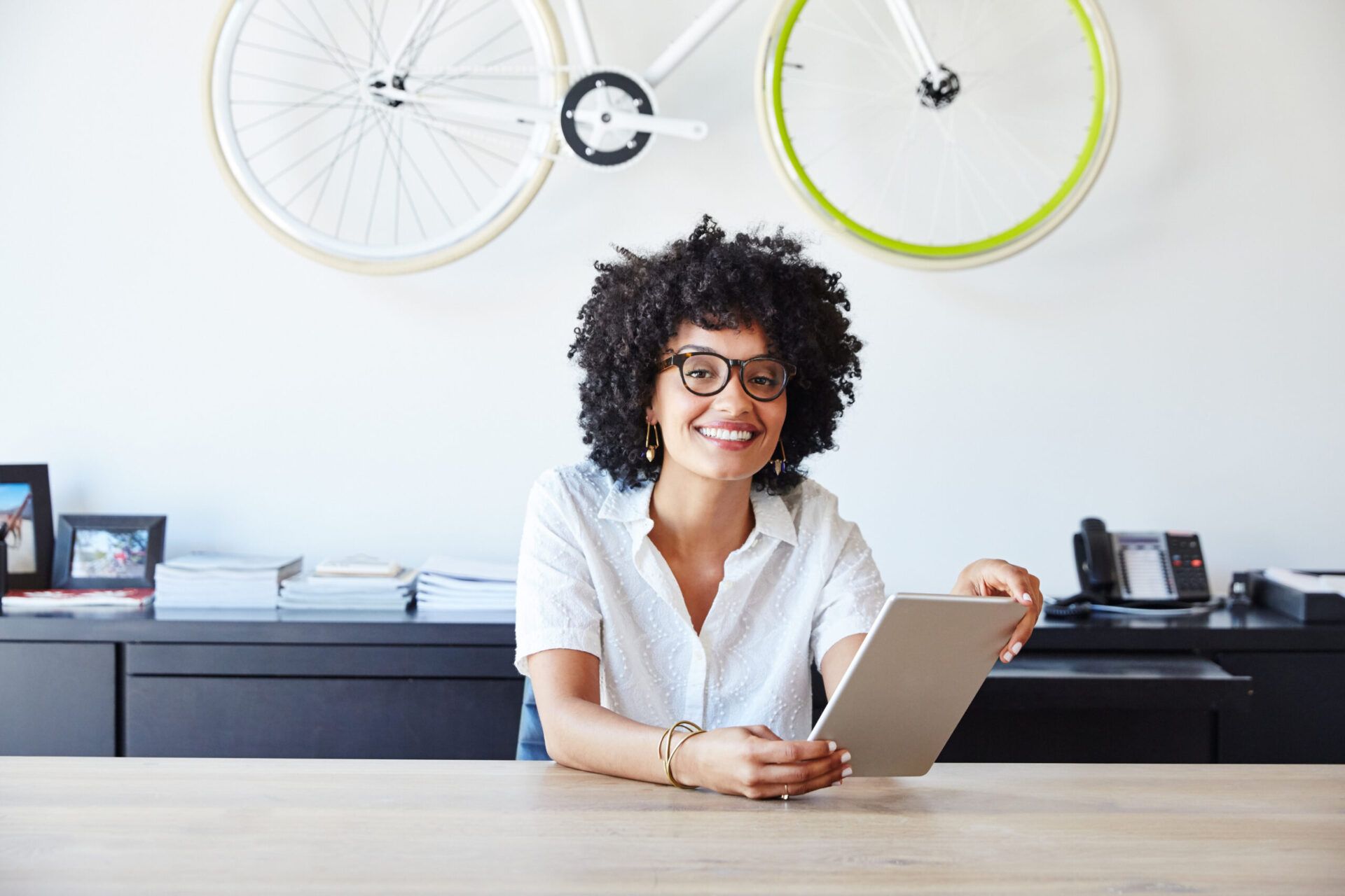 Woman in glasses and a white shirt holding a tablet and smiling in front of a decorative hanging bicycle
