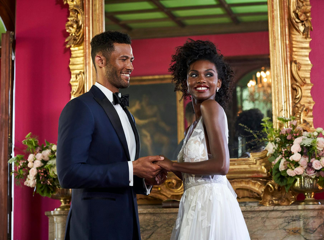 A couple holding hands smiles joyfully, dressed in formal attire. The elegant room has pink walls, a large ornate mirror, and floral arrangements.