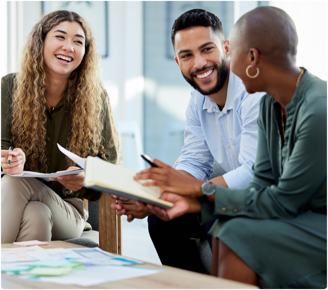 Three people are sitting together, smiling and engaged in discussion. They hold notebooks and papers in a brightly lit, modern office setting.