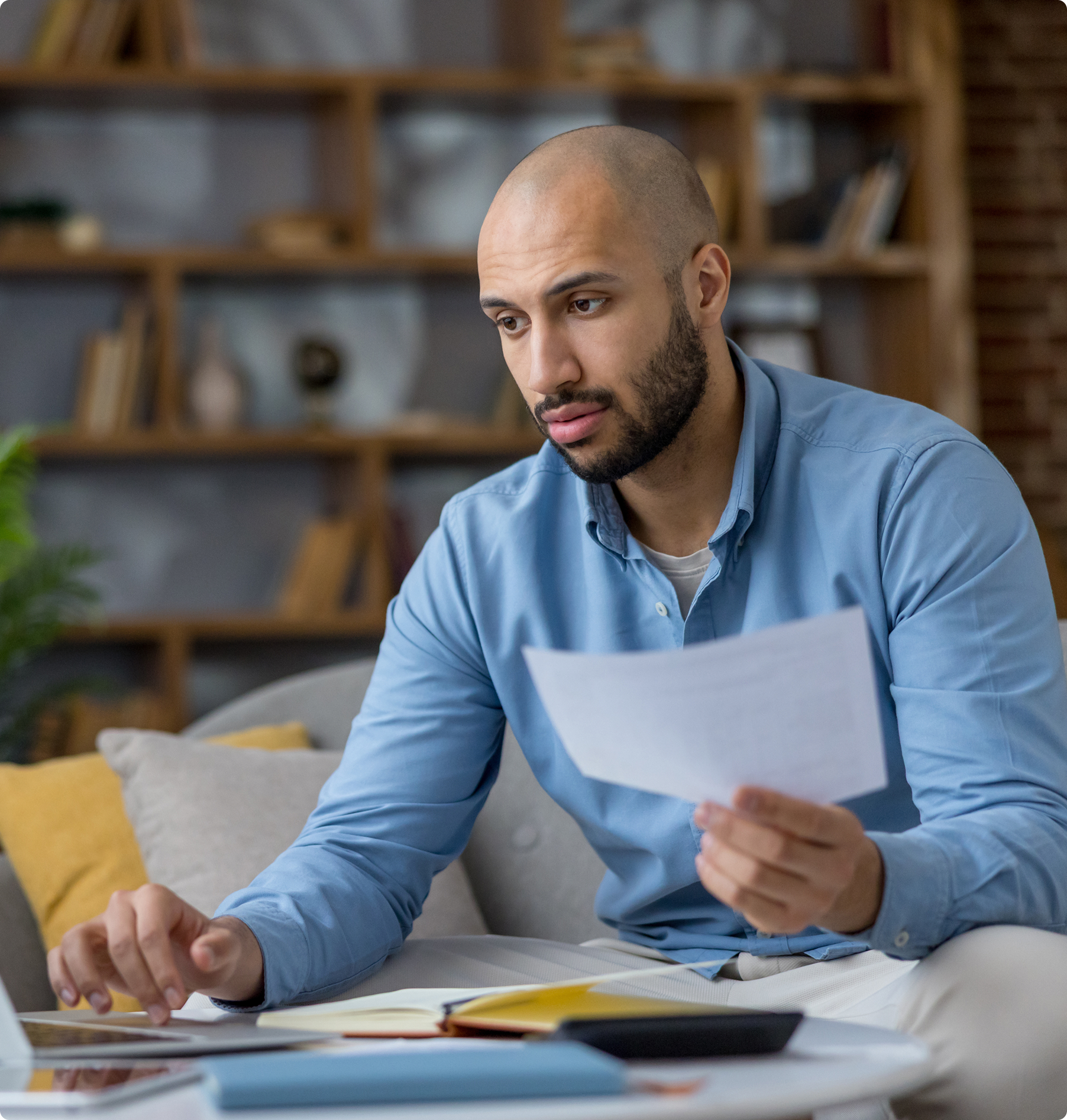 A man in a blue shirt sits on a sofa, focusing intently on a paper in hand. Nearby is a laptop and notebooks. The setting is a cozy room with a bookshelf.