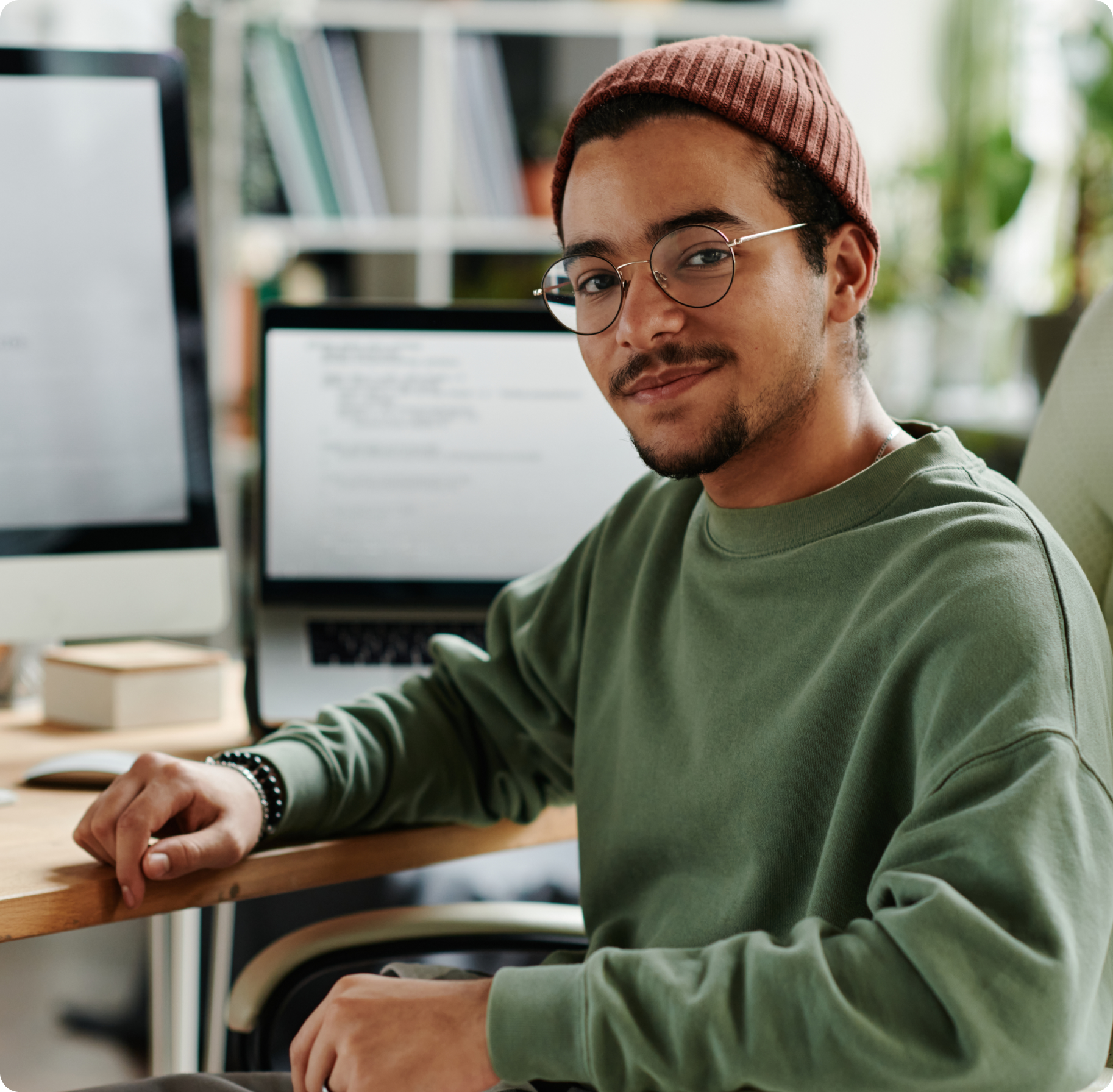 Man in glasses and a beanie smiles at a desk with a computer. The background shows a blurred interior with books and plants, conveying a cozy workspace.