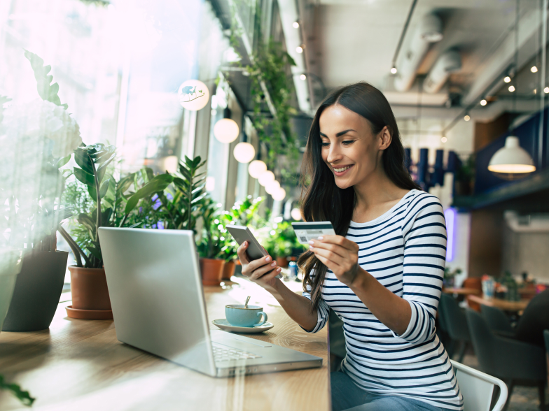 A millennial woman shops online at a cafe, holding her phone and credit card with a laptop open in front of her