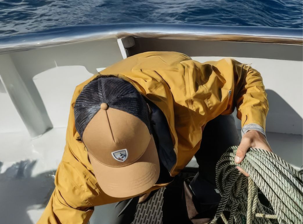 A person in a yellow jacket and cap organizes ropes on a boat deck against a backdrop of calm ocean and distant islands, conveying a sense of adventure.