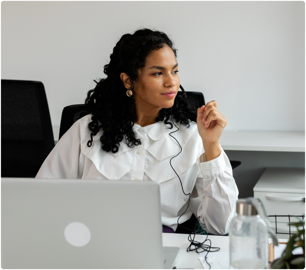 A woman with curly hair and gold earrings sits at a desk, wearing a white blouse and headset. She's gazing thoughtfully to the side.
