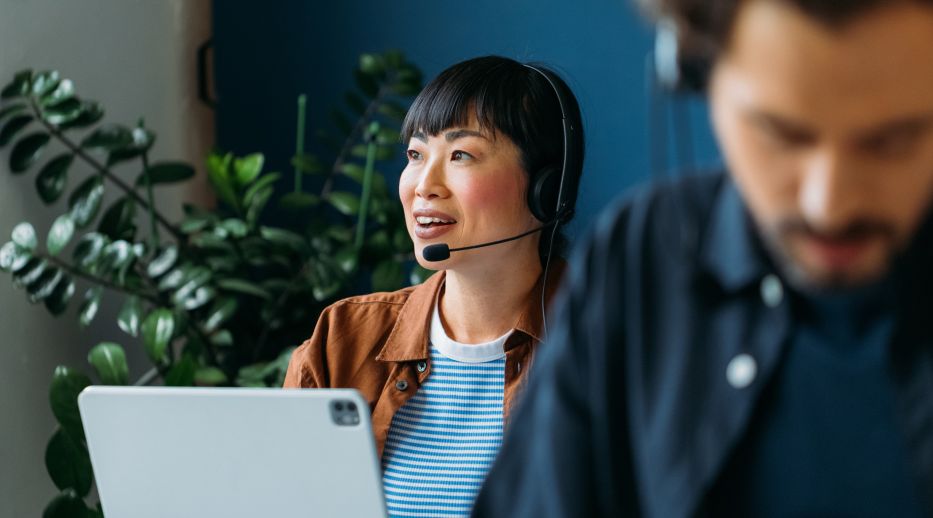 A woman with a headset smiles while working on a laptop in a bright, casual office setting. A plant and another person with a headset are in the background.