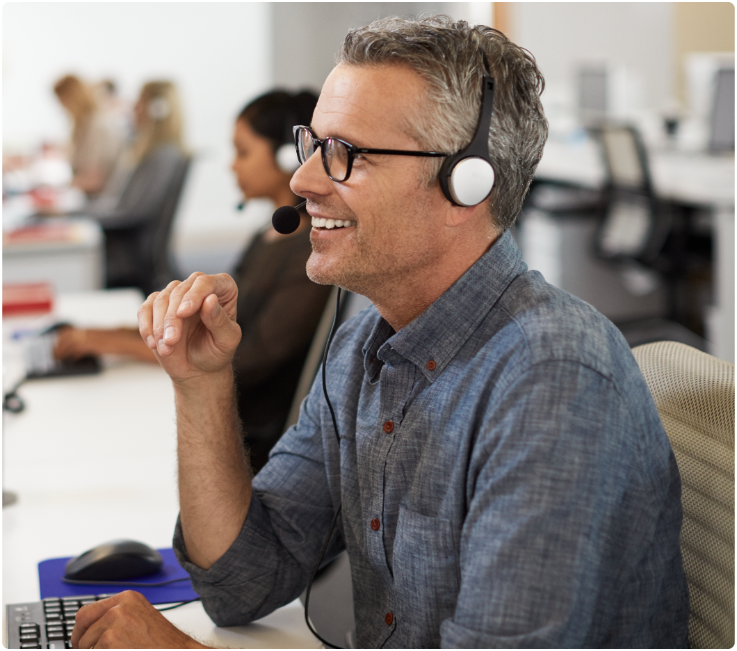 A man with glasses and gray hair, wearing a headset, smiles while working at a computer in a bright office. Colleagues are in the background.