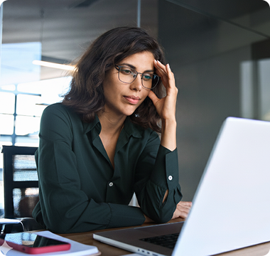 A man in a blue shirt sits at a desk with a laptop, looking stressed, holding his forehead. The room has soft lighting and a blurred view outside.