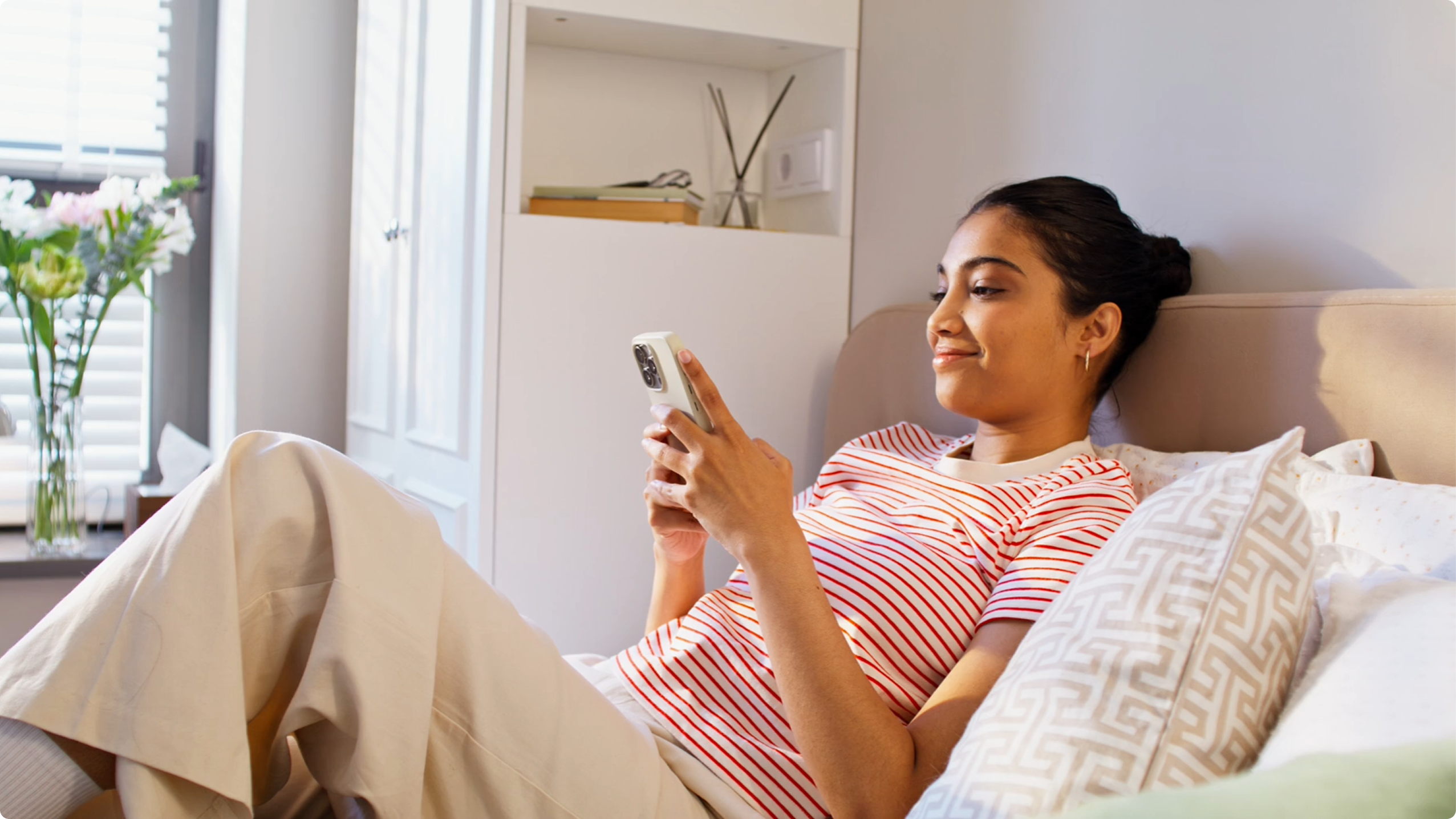 A woman in a striped shirt relaxes on a couch, smiling while looking at her phone. Sunlight streams in through a window with a vase of flowers nearby.