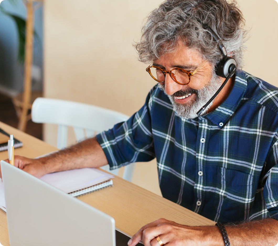 Smiling man with gray hair and glasses wearing a headset, sitting at a desk with a laptop and notepad. He appears engaged and content.