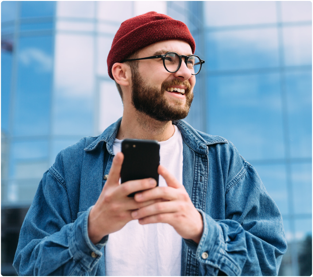 A bearded man wearing a red beanie and glasses smiles while holding a smartphone. He is outdoors, in front of a modern glass building, conveying a cheerful mood.