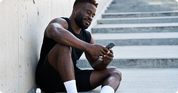 Man in athletic wear sits on outdoor steps, smiling while looking at his smartphone. A water bottle is beside him, conveying a relaxed, post-workout vibe.