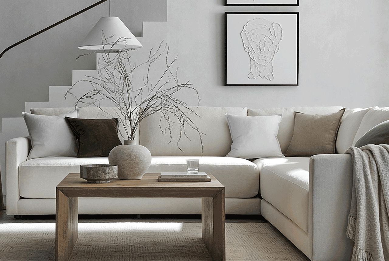 A minimalist living room features a white sectional sofa adorned with neutral cushions. A wooden coffee table holds a ceramic vase with twigs, under soft art prints.