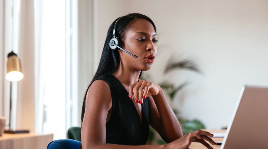 A woman wearing a black top and a headset sits at a desk, intently working on a laptop. A lamp and plant are in the blurred background, conveying a professional and focused environment.