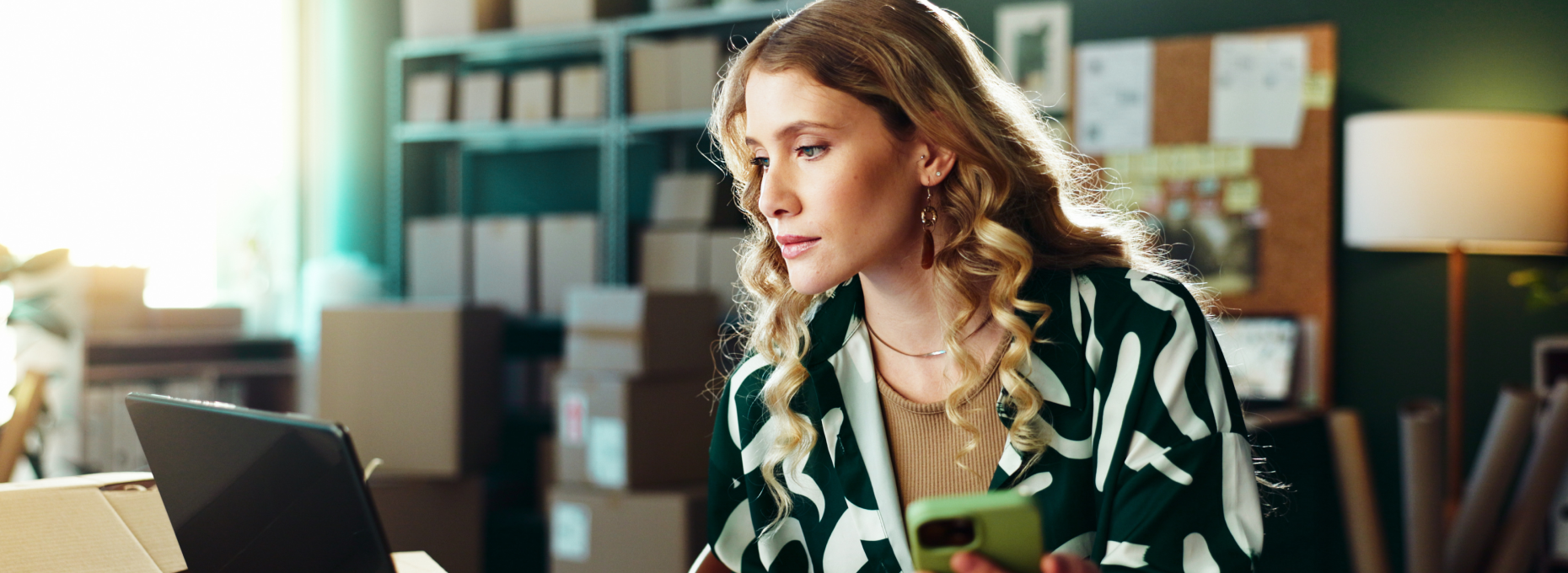 Small business owner checking her phone and laptop in a home office stocked with shipping boxes