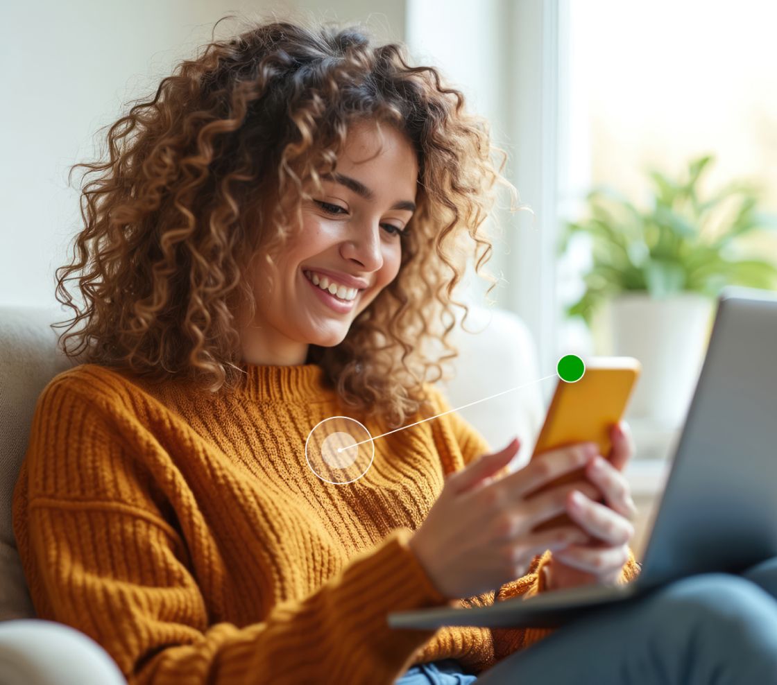 Smiling woman in a cozy orange sweater using a smartphone while relaxing. A laptop is nearby, with a vibrant plant in the background. Bright, cheerful mood.