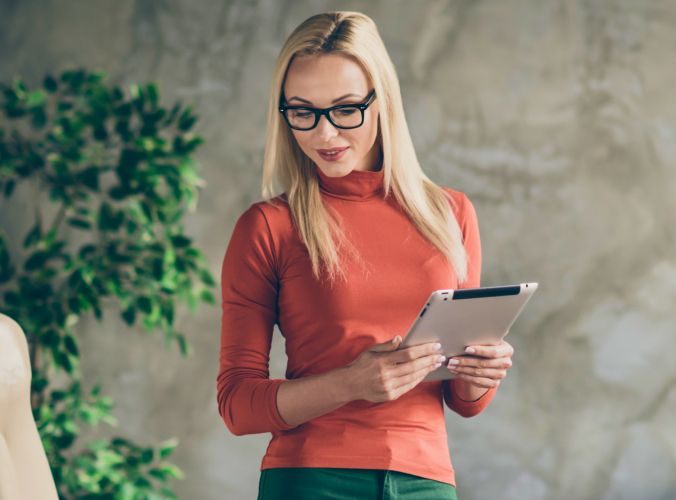 A woman with long blonde hair and glasses stands holding a tablet. She wears an orange turtleneck, exuding a relaxed, professional vibe.