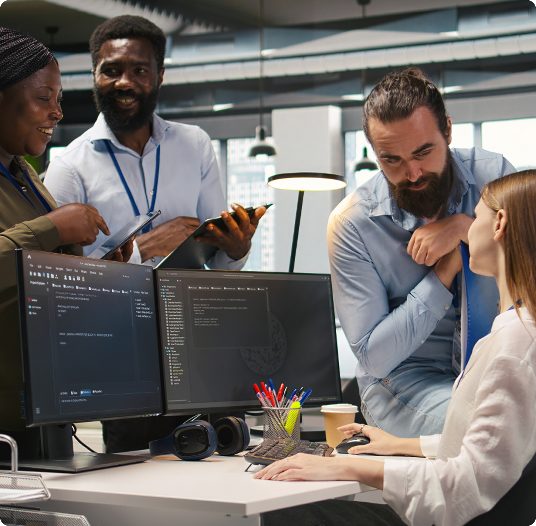 A diverse group of four colleagues discusses coding at a modern office. They appear engaged and collaborative, with computers and notes on the desk.