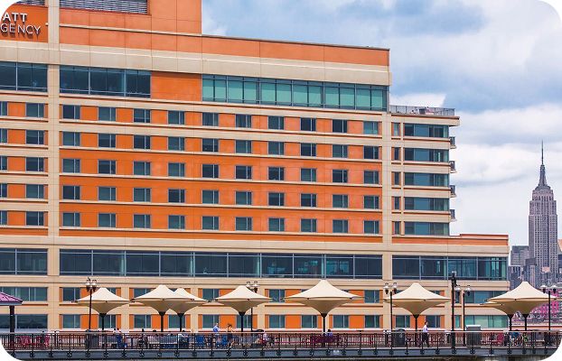 Large orange hotel building under a cloudy sky, featuring rows of windows. In front, beige umbrellas line a promenade. The Empire State Building is visible in the background, adding a touch of New York City charm.