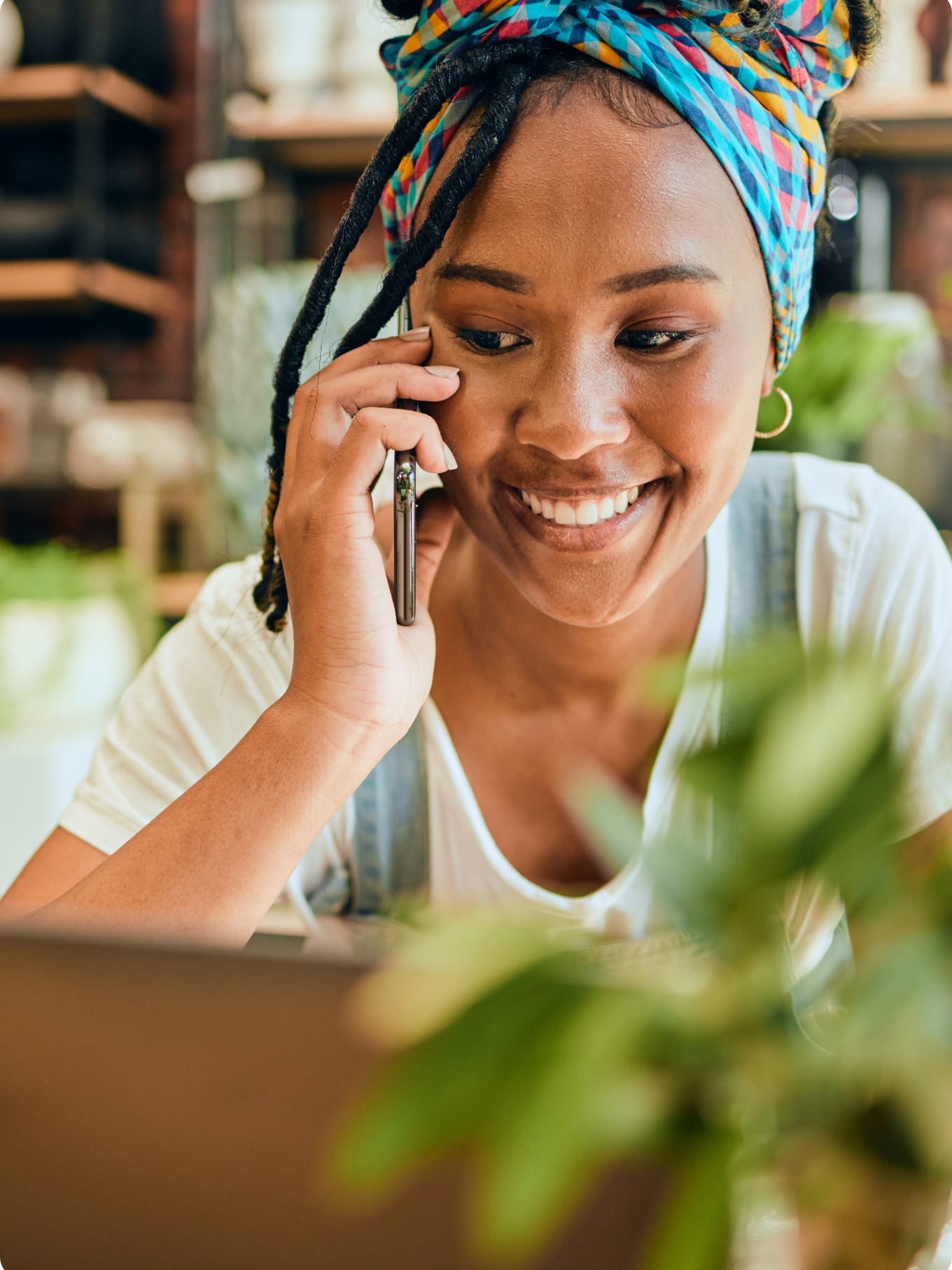 Smiling woman talking on a phone, wearing a colorful headscarf, with plants blurred in the foreground, conveying a warm, cheerful atmosphere.