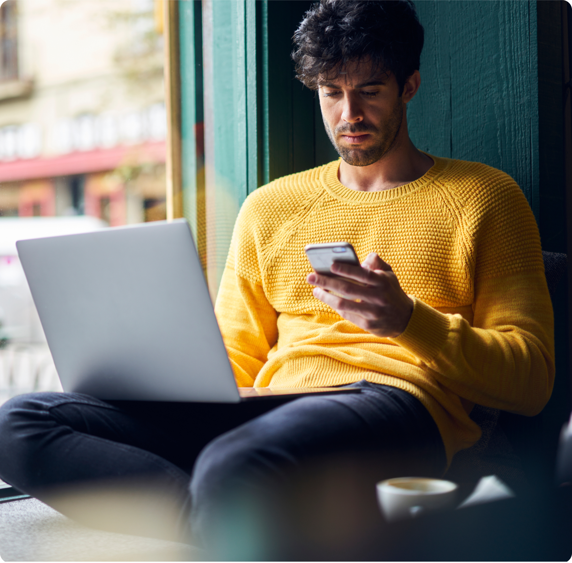 A man in a yellow sweater sits by a window, focused on his smartphone. He has a laptop on his lap and a coffee cup beside him, conveying a relaxed, studious mood.