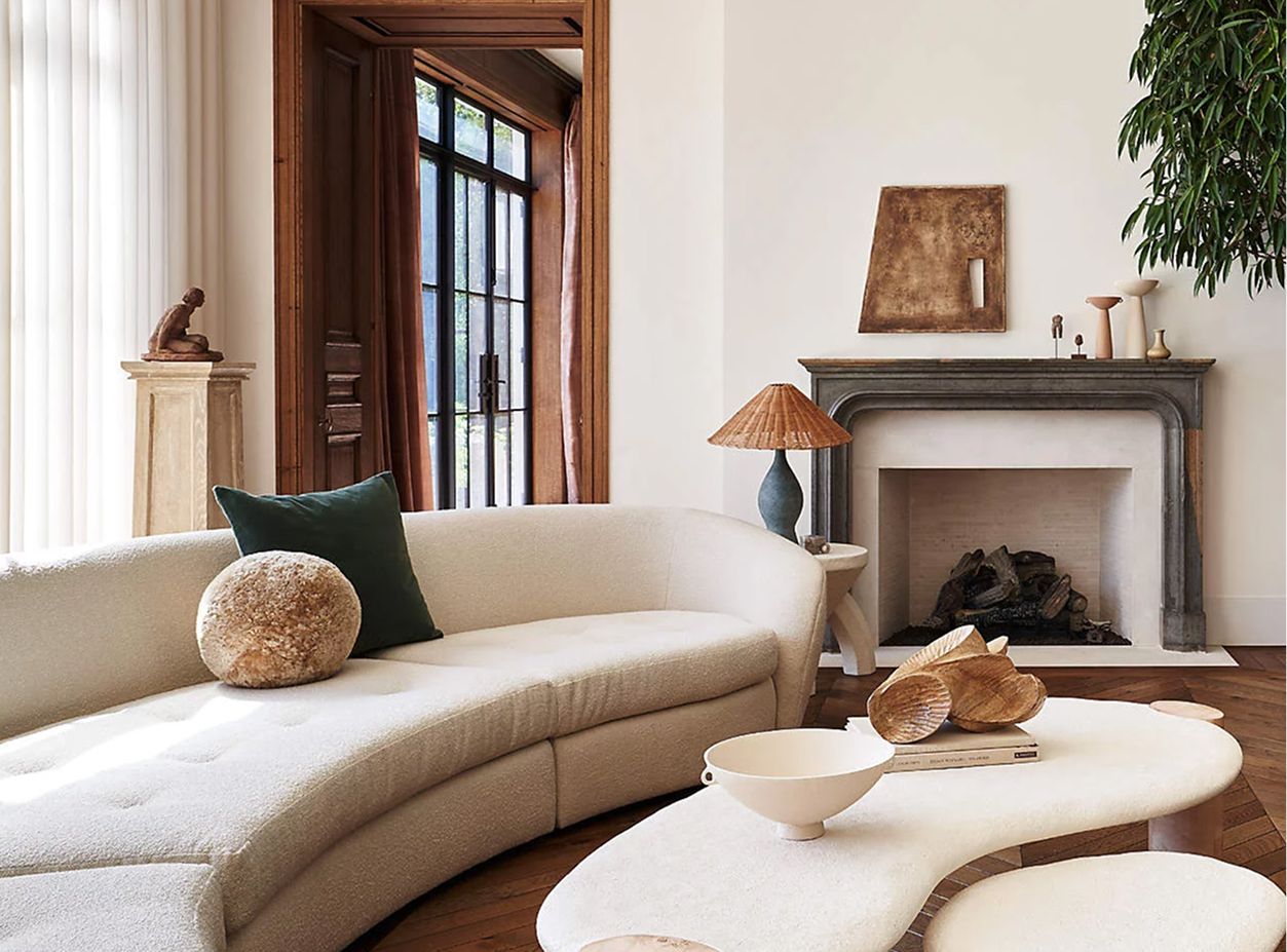Contemporary living room with a curved beige sofa, wooden coffee table, and decorative items. Warm tones, relaxed atmosphere, fireplace, large window.