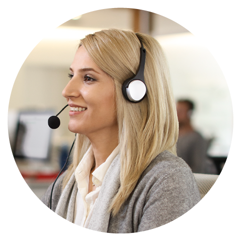 A woman with blonde hair wearing a headset smiles while sitting at a desk in an office environment, conveying a professional and friendly tone.