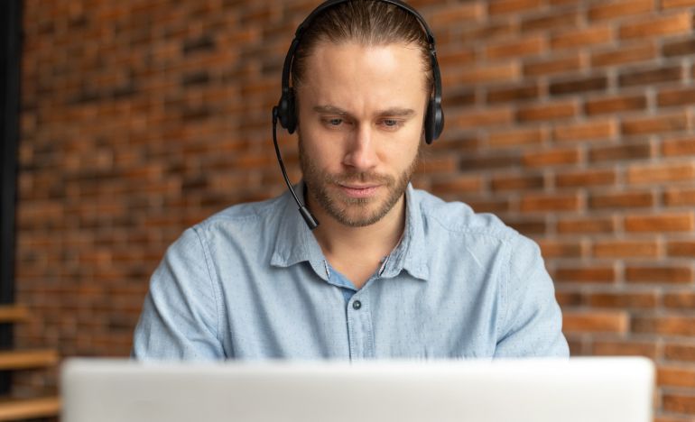 Bearded man with a phone headset looking at a laptop
