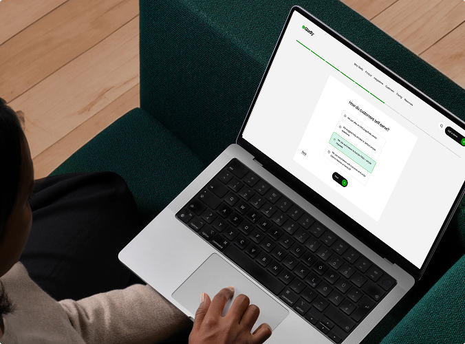 A person sits on a dark green couch using a laptop displaying a survey form. The wooden floor and relaxed posture create a focused yet casual atmosphere.