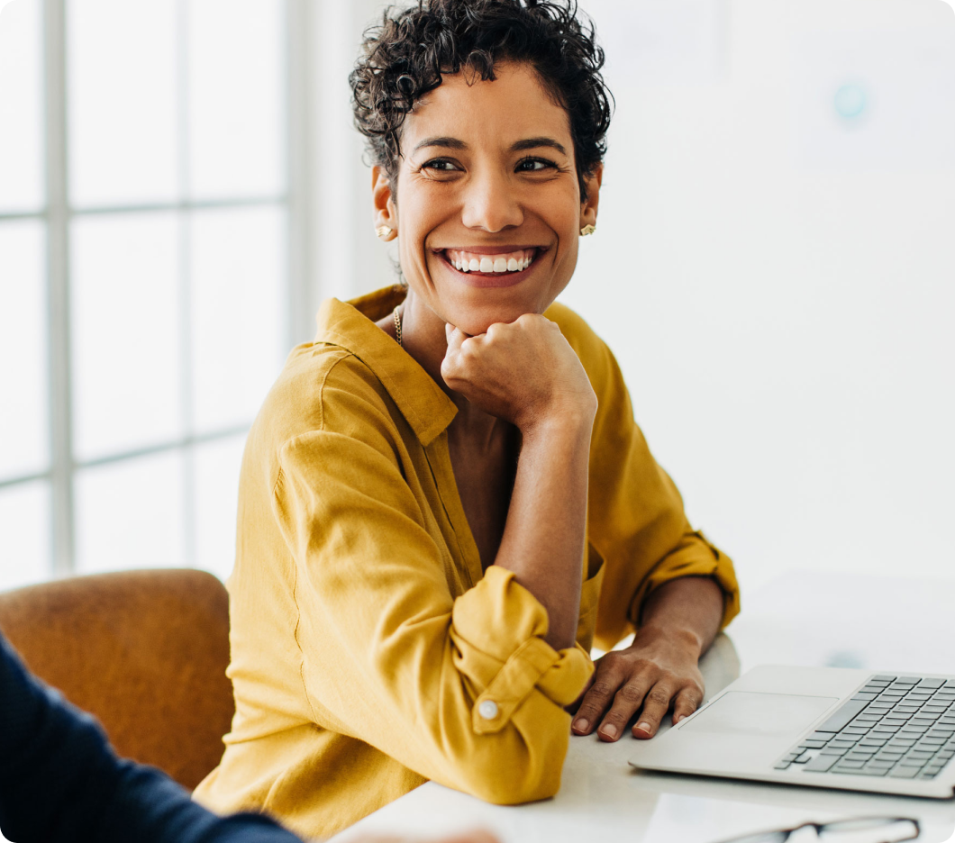 Smiling person in a yellow shirt, seated at a desk with a laptop, looking to the side. The image conveys warmth and friendliness in a bright room.