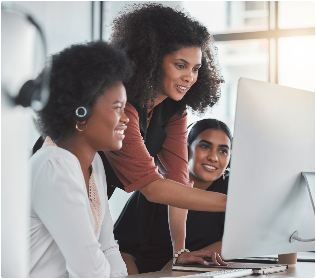 Three women in an office setting collaborate around a computer. One points at the screen, while another smiles, wearing a headset. Bright, focused atmosphere.