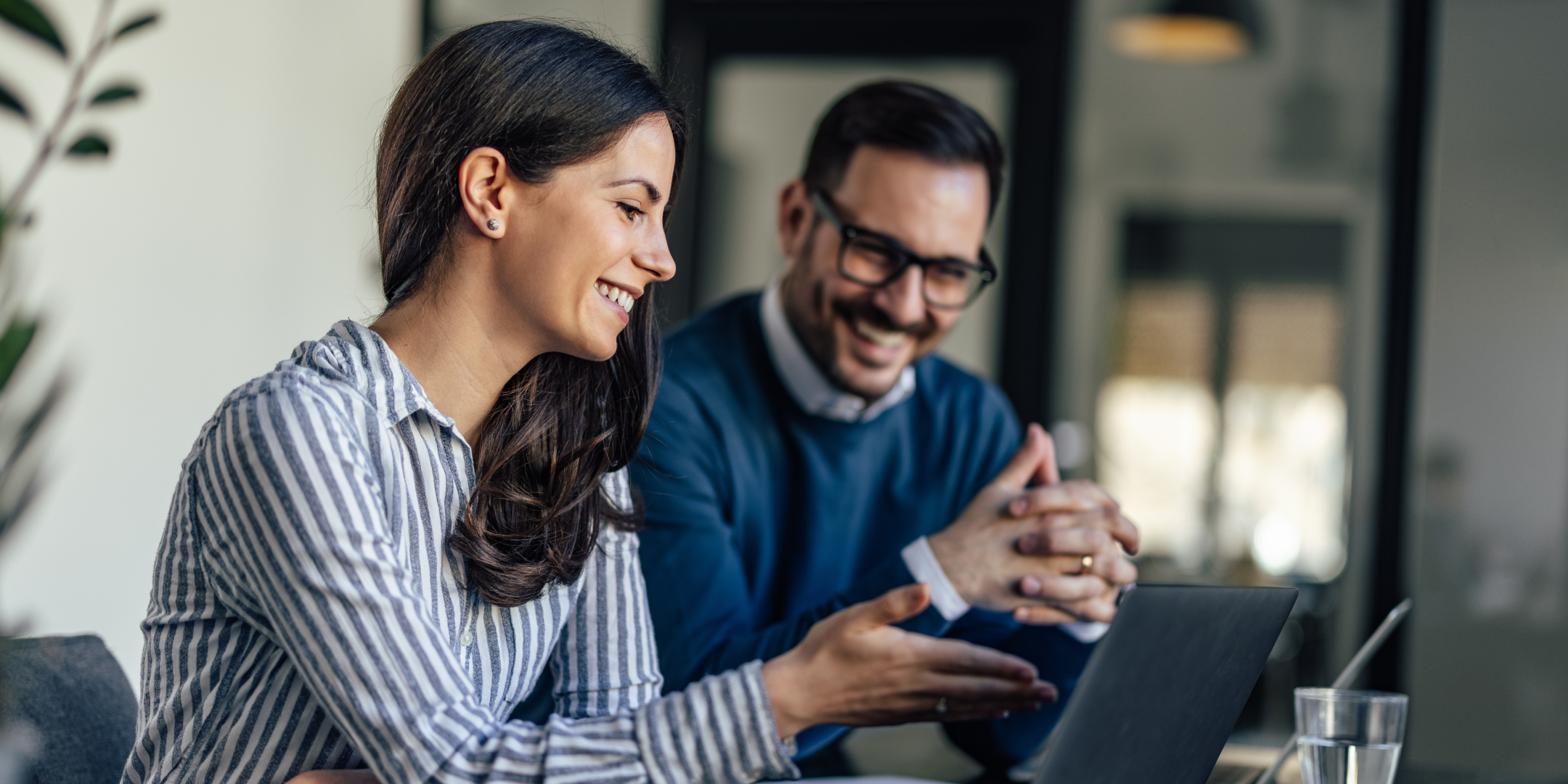 A man and woman are smiling and engaged in a discussion while looking at a laptop in a modern office. The atmosphere is collaborative and positive.