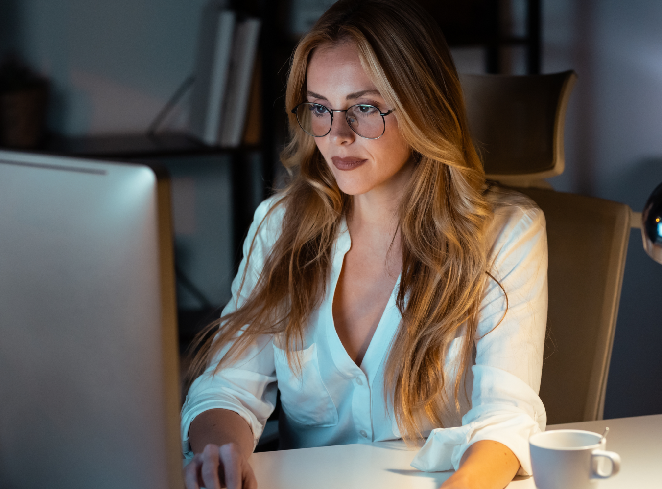 A woman with long hair and glasses is focused on a computer in a dimly lit office. A cup sits nearby on the desk, conveying a quiet, studious atmosphere.