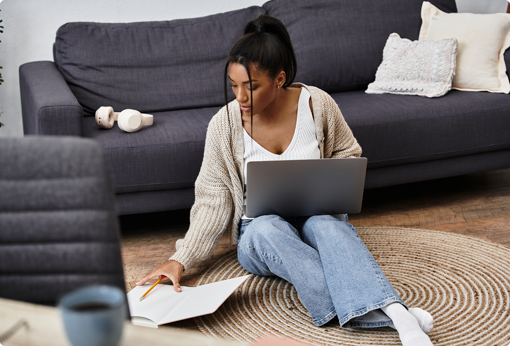 Young woman with a laptop sits on the floor, focused on taking notes in an open book. Nearby, a sofa, pillows, and headphones create a cozy, calm atmosphere.