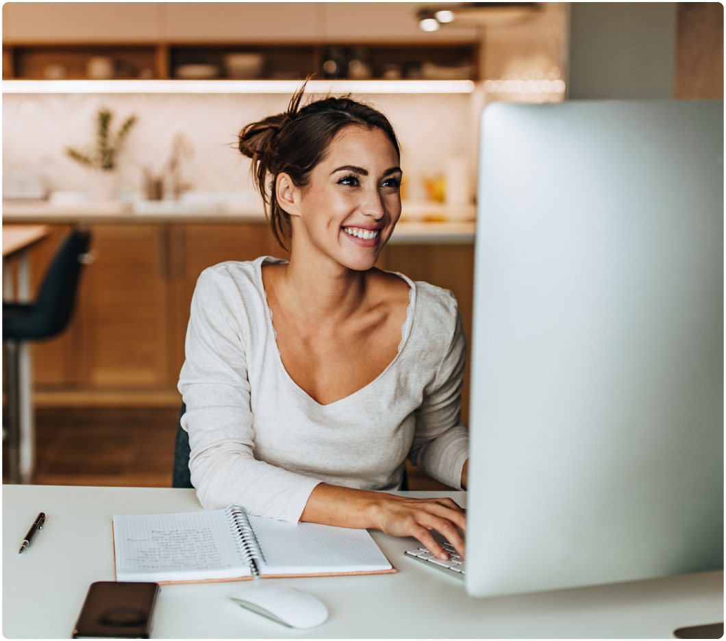 Smiling woman working on a computer at a table with a notebook and phone, set in a cozy kitchen with warm lighting. Tone is cheerful and productive.
