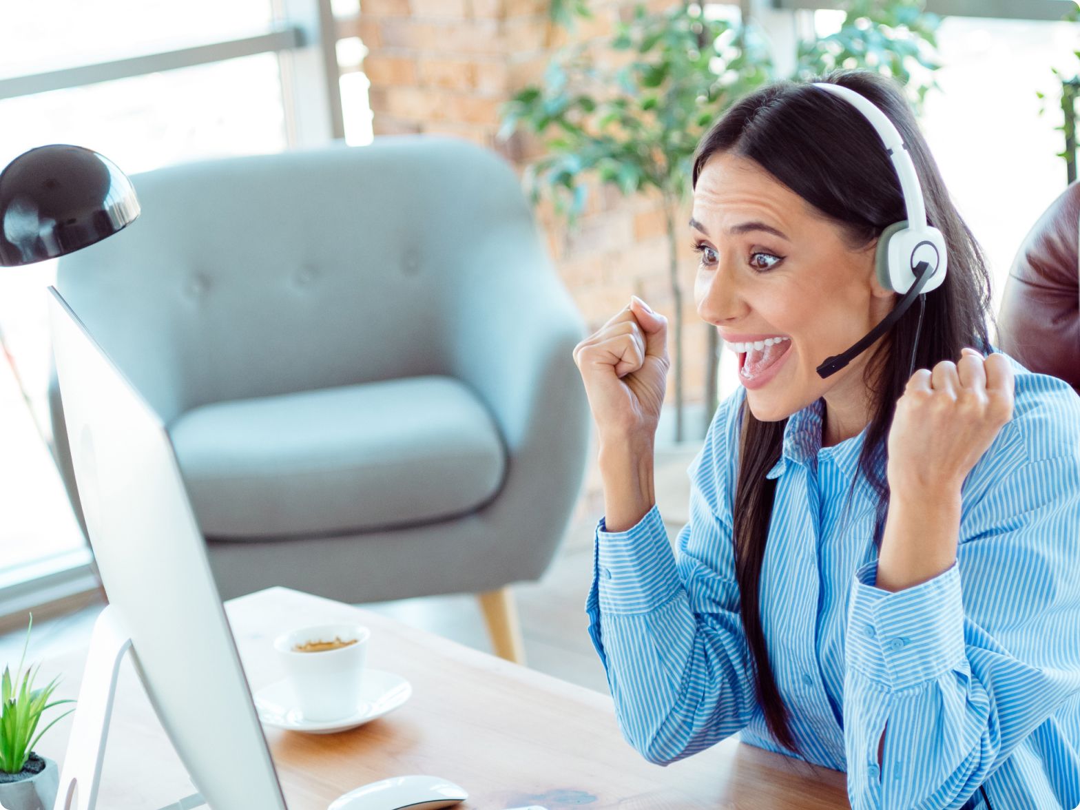A woman in a blue striped shirt excitedly raises her fists, wearing a headset in front of a computer. Her expression conveys enthusiasm and success.