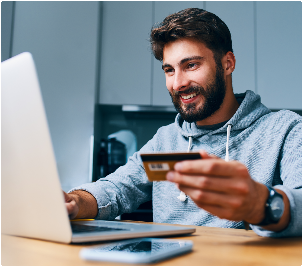 A smiling man in a gray hoodie holds a credit card while using a laptop at a wooden table. The scene is well-lit, conveying a sense of ease.