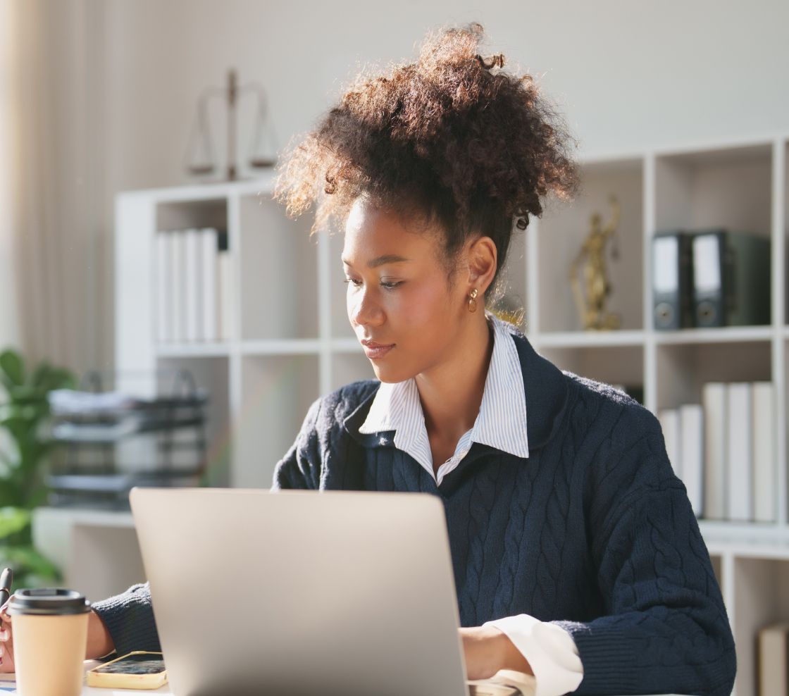 A focused woman in a navy sweater works at a laptop in a bright office, with bookshelves behind her and a coffee cup nearby, conveying concentration.