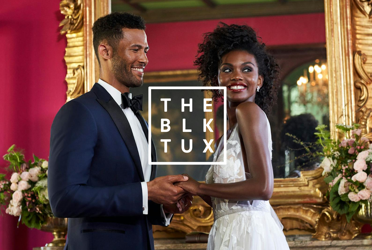 A couple holding hands smiles joyfully, dressed in formal attire. The elegant room has pink walls, a large ornate mirror, and floral arrangements.