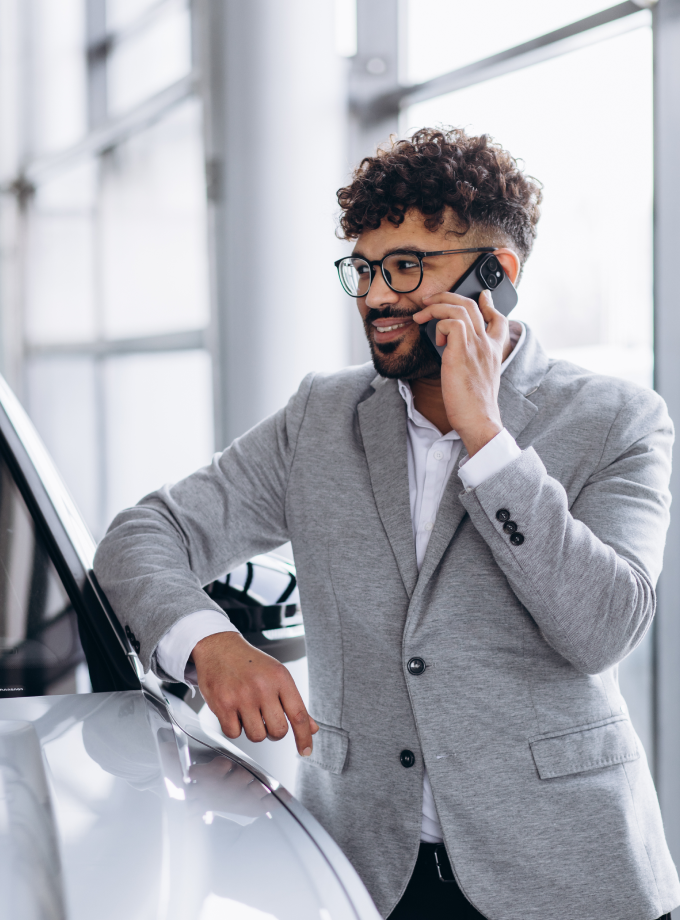 A man in a gray suit and glasses, with curly hair, smiles while talking on a phone. He's leaning against a car in a bright, modern setting.