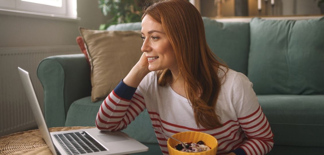 A woman with red hair wearing a striped shirt sits on the floor, smiling at a laptop. She's holding a bowl of yogurt. The room feels cozy and relaxed.