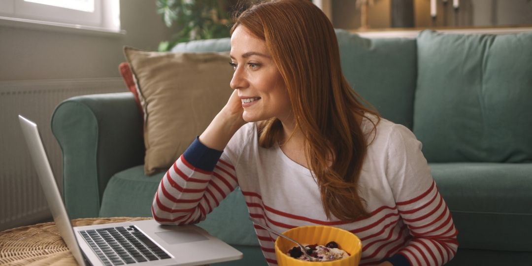 A woman with red hair wearing a striped shirt sits on the floor, smiling at a laptop. She's holding a bowl of yogurt. The room feels cozy and relaxed.