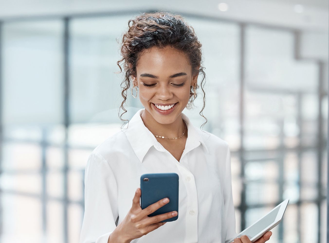 A woman in a white blouse smiles while looking at her smartphone. She holds a tablet, standing in a bright, modern office. The mood is cheerful and professional.