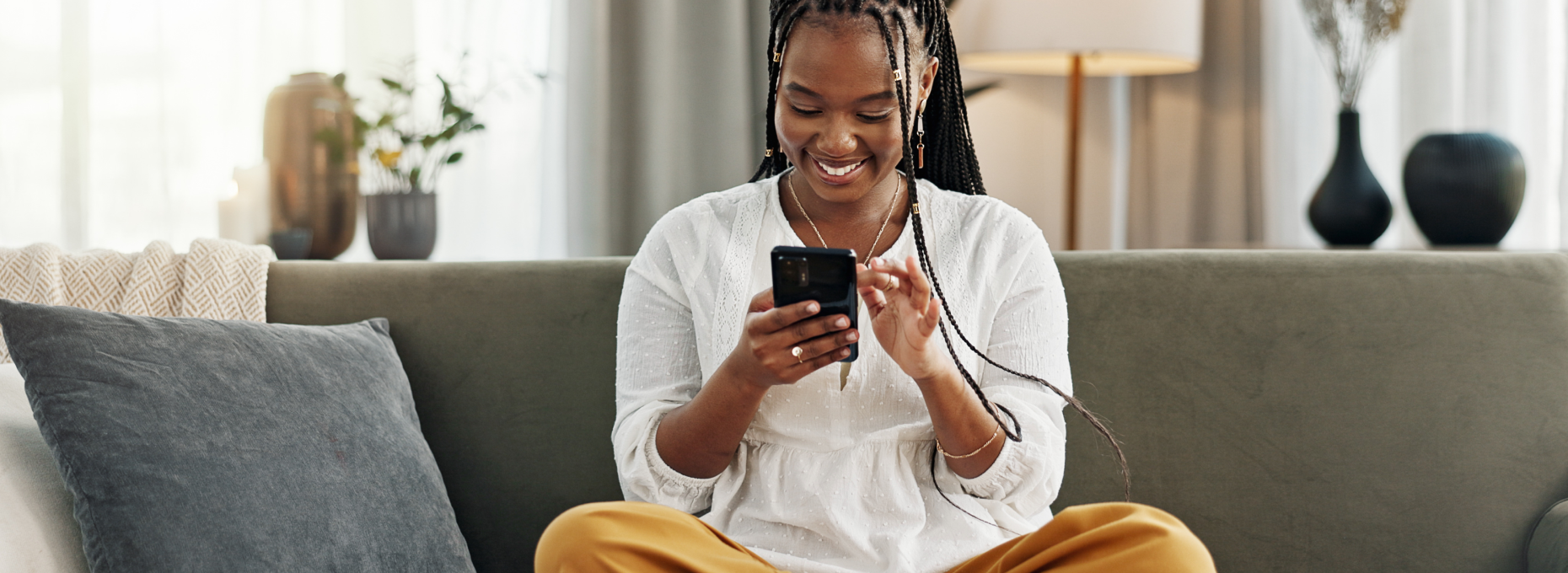Woman sitting cross-legged on a gray couch at home, smiling as she shops on her phone