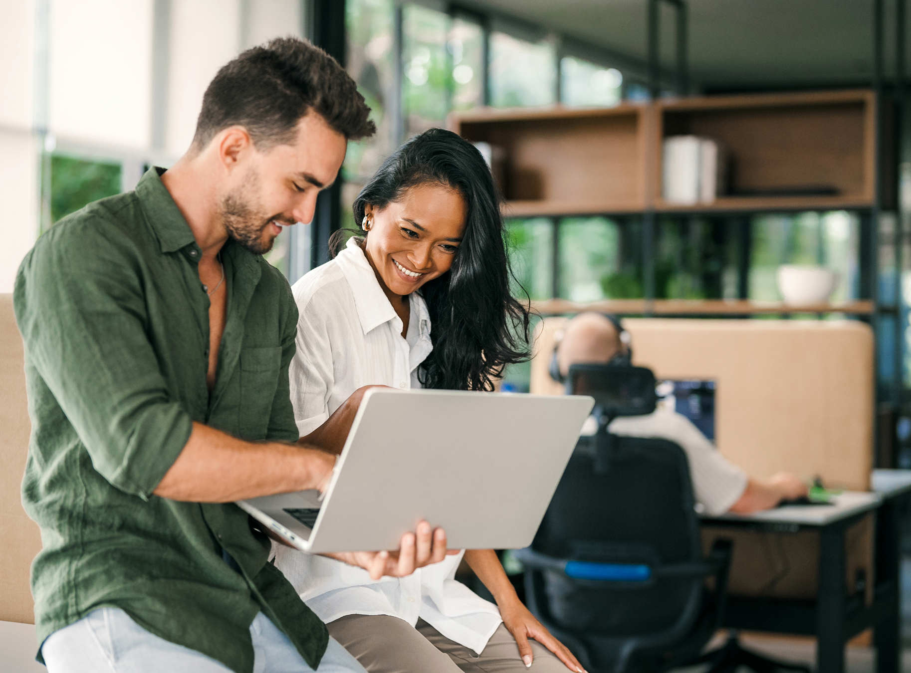 A smiling man and woman collaborate on a laptop in a modern office, with large windows in the background, conveying teamwork and positivity.