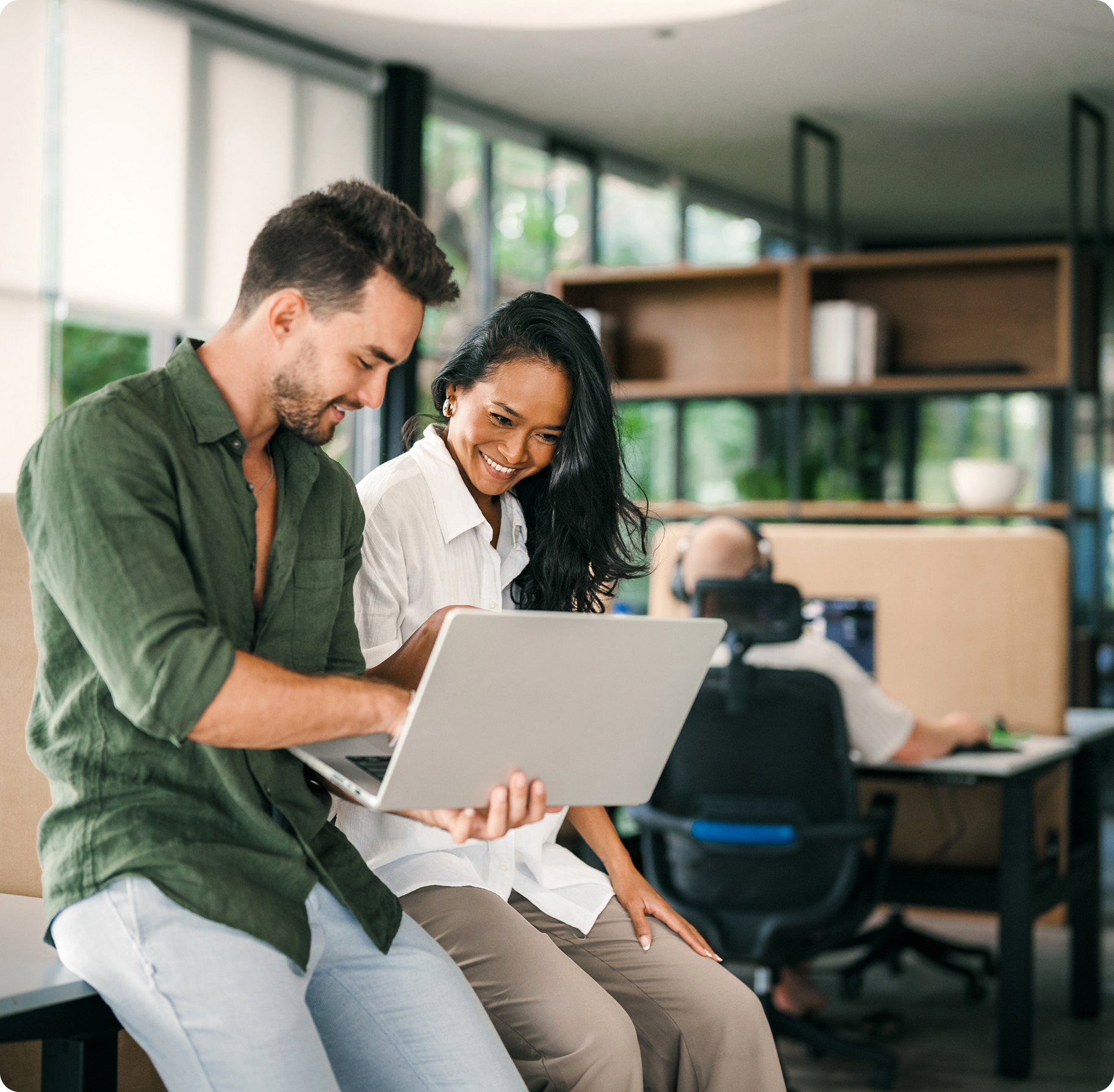 A smiling man and woman collaborate on a laptop in a modern office, with large windows in the background, conveying teamwork and positivity.