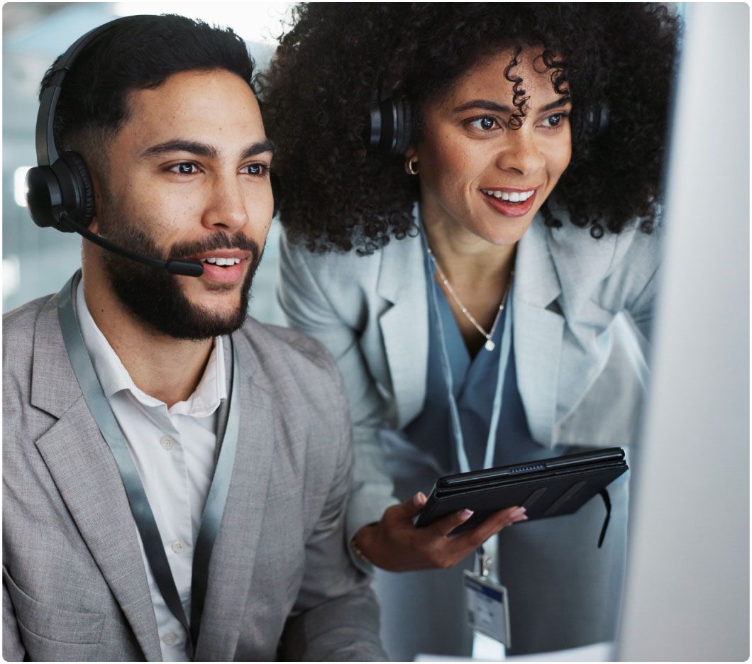 Two colleagues wearing headsets are collaborating at a computer in an office. Both are smiling, suggesting a positive and engaging work environment.