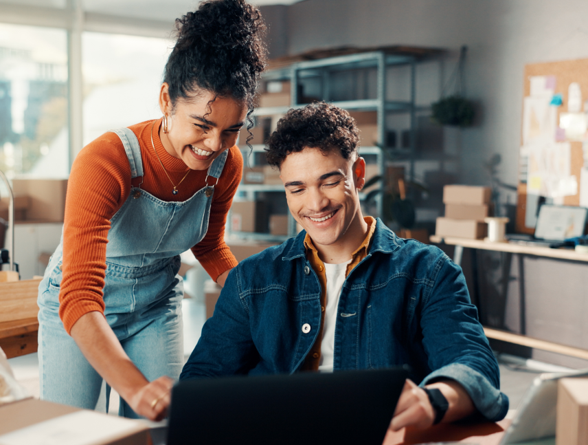 Two coworkers smiling while they review something on a laptop together in a small ecommerce stockroom with shelves and shipping boxes