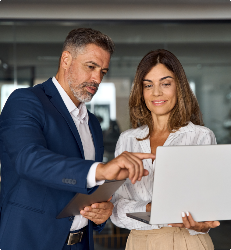 Man in a blue suit gestures at a laptop screen held by a woman in a white blouse, standing in a modern office. They appear focused and collaborative.
