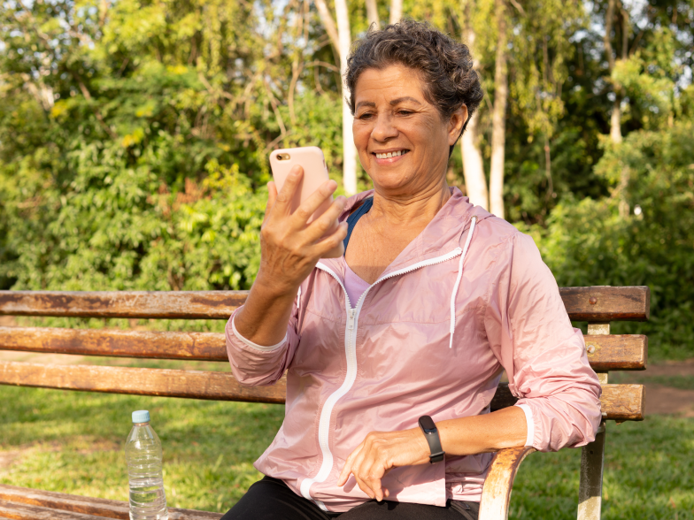 Smiling woman in a pink windbreaker on a park bench, reading a reply from a brand on her phone